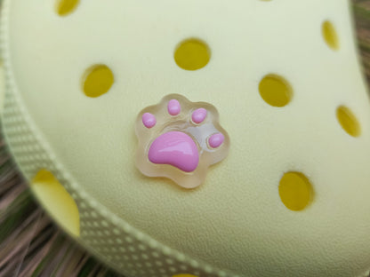 Glow-in-the-dark pastel paw print charms in pink, yellow, blue, and green, attached to a yellow shoe. The charms have a soft glow effect and add a playful animal-inspired look.
