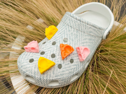 Close-up of a pair of light blue shoes decorated with colorful 3D-printed cheese wedge charms in shades of yellow, orange, and pink. The cheese charms feature realistic holes and a playful, quirky design, adding a fun pop of color to the footwear.