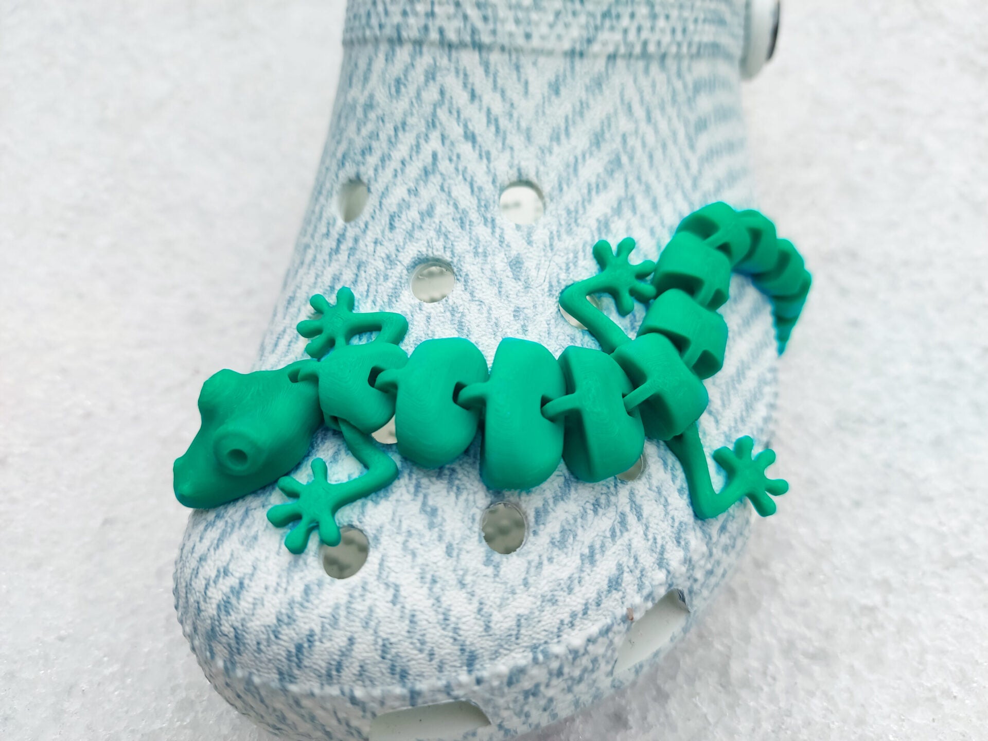 Three colorful 3D printed flexi lizard charms in red, green, and yellow, clipped onto blue-patterned shoes. The articulated lizards appear lifelike with detailed feet and movable segments, set against a snowy white backdrop.
