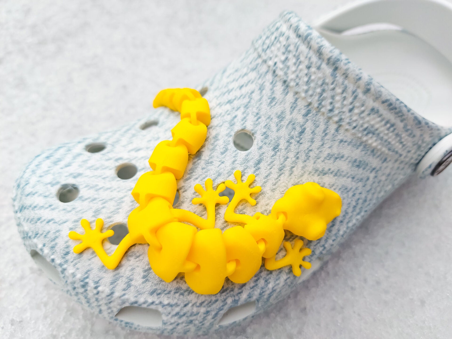 Three colorful 3D printed flexi lizard charms in red, green, and yellow, clipped onto blue-patterned shoes. The articulated lizards appear lifelike with detailed feet and movable segments, set against a snowy white backdrop.