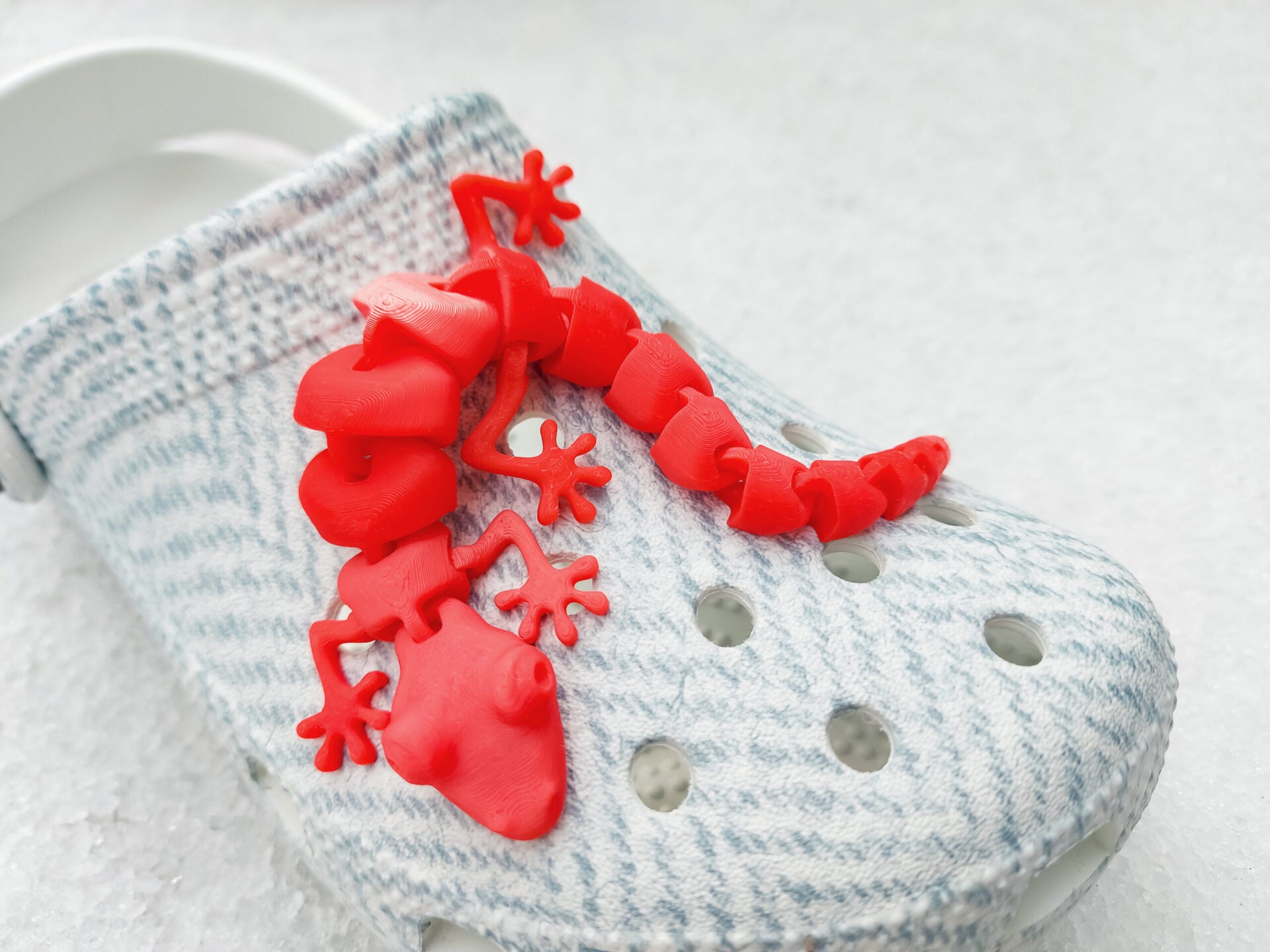 Three colorful 3D printed flexi lizard charms in red, green, and yellow, clipped onto blue-patterned shoes. The articulated lizards appear lifelike with detailed feet and movable segments, set against a snowy white backdrop.