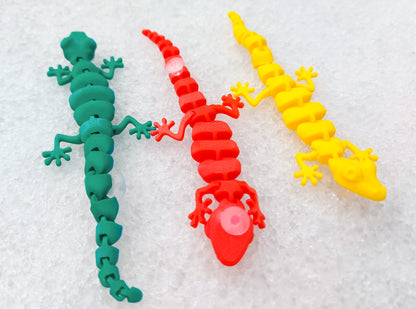 Three colorful 3D printed flexi lizard charms in red, green, and yellow, clipped onto blue-patterned shoes. The articulated lizards appear lifelike with detailed feet and movable segments, set against a snowy white backdrop.