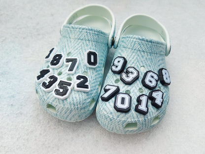 A pair of blue-patterned shoes featuring custom 3D-printed number charms, showcasing the digits 0 through 9 in sleek black and white colors. Each charm is securely clipped into the shoe holes, standing out with a bold varsity-inspired style.