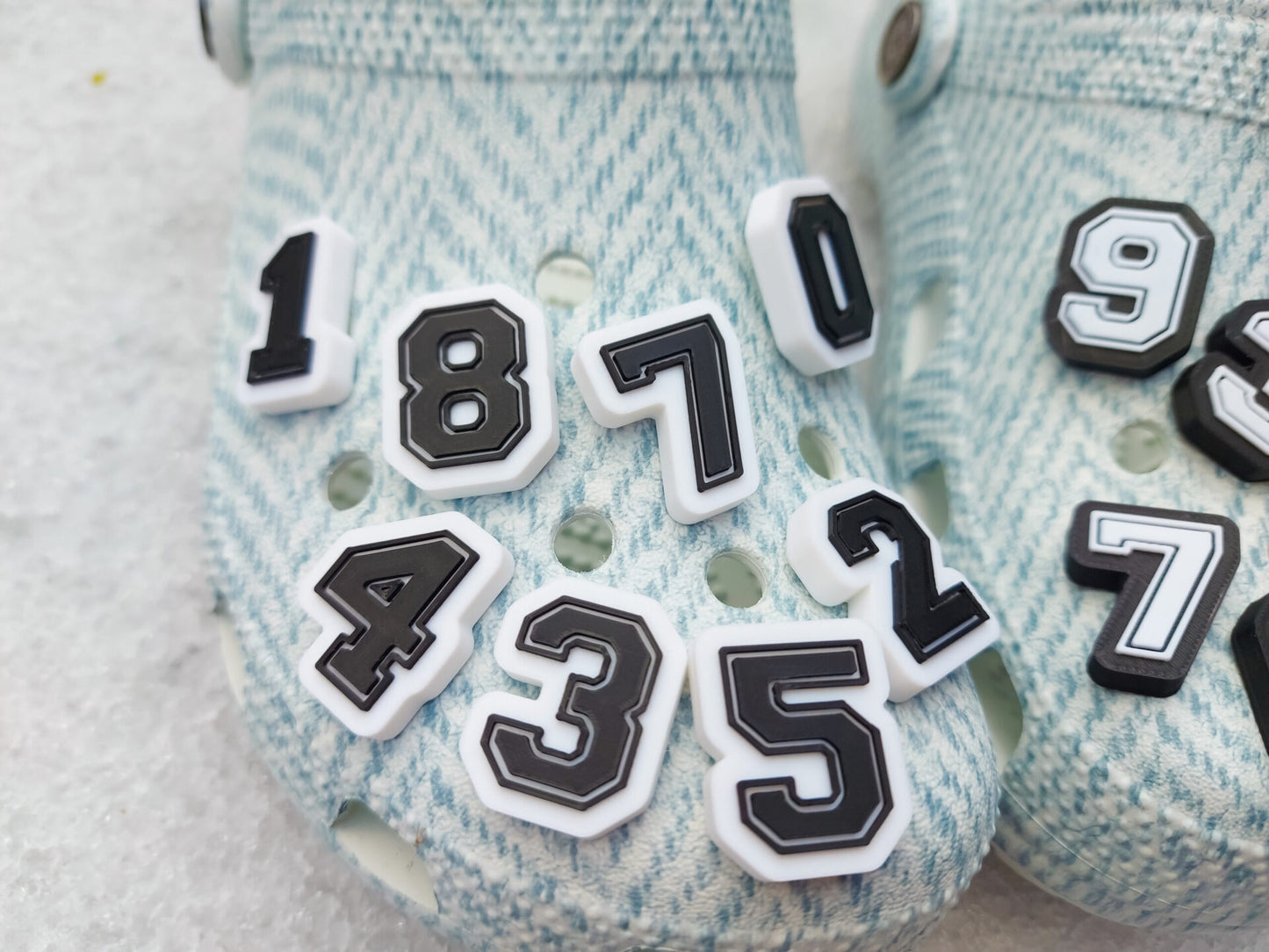 A pair of blue-patterned shoes featuring custom 3D-printed number charms, showcasing the digits 0 through 9 in sleek black and white colors. Each charm is securely clipped into the shoe holes, standing out with a bold varsity-inspired style.