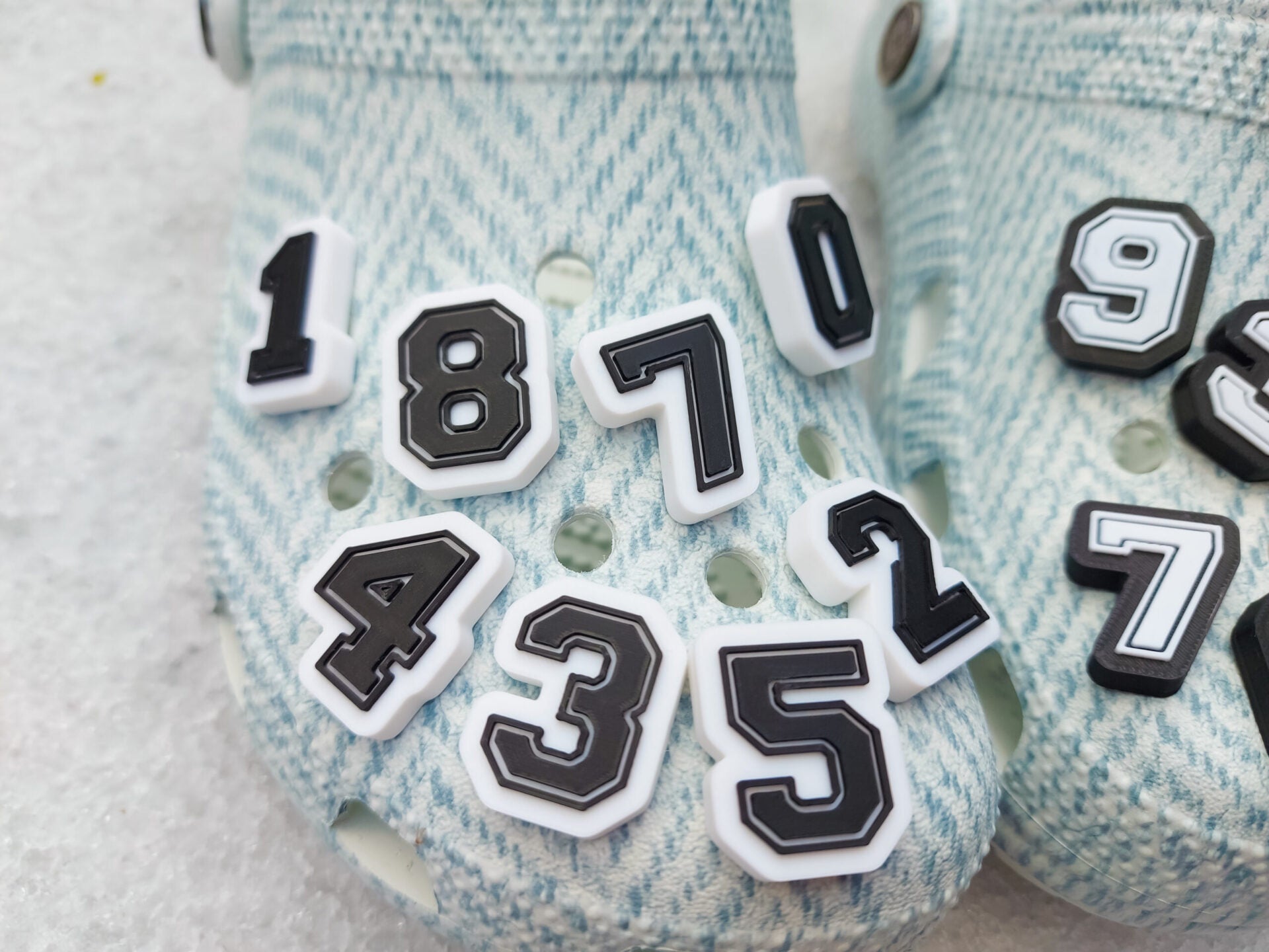 A pair of blue-patterned shoes featuring custom 3D-printed number charms, showcasing the digits 0 through 9 in sleek black and white colors. Each charm is securely clipped into the shoe holes, standing out with a bold varsity-inspired style.