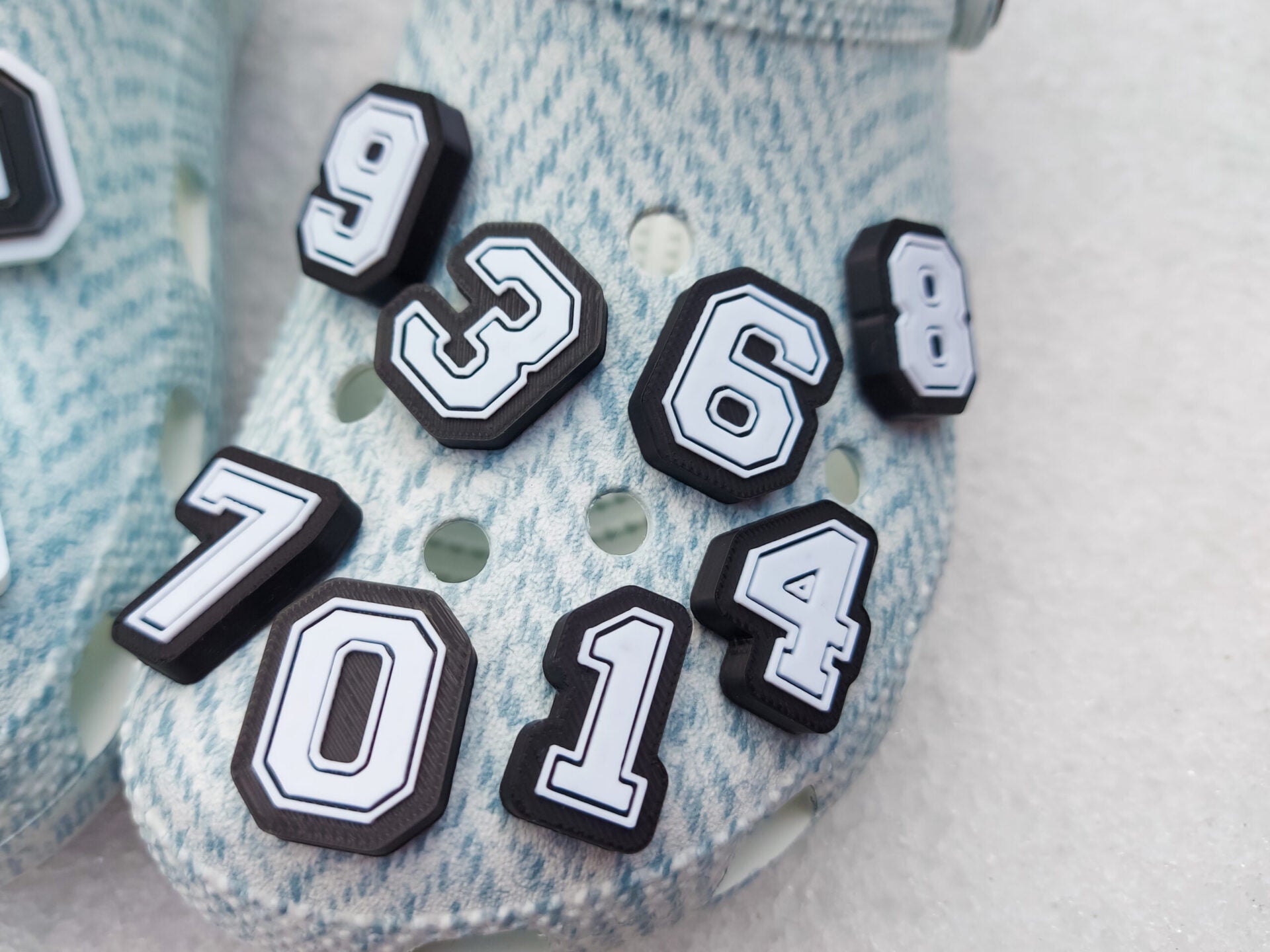 A pair of blue-patterned shoes featuring custom 3D-printed number charms, showcasing the digits 0 through 9 in sleek black and white colors. Each charm is securely clipped into the shoe holes, standing out with a bold varsity-inspired style.