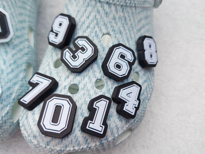 A pair of blue-patterned shoes featuring custom 3D-printed number charms, showcasing the digits 0 through 9 in sleek black and white colors. Each charm is securely clipped into the shoe holes, standing out with a bold varsity-inspired style.