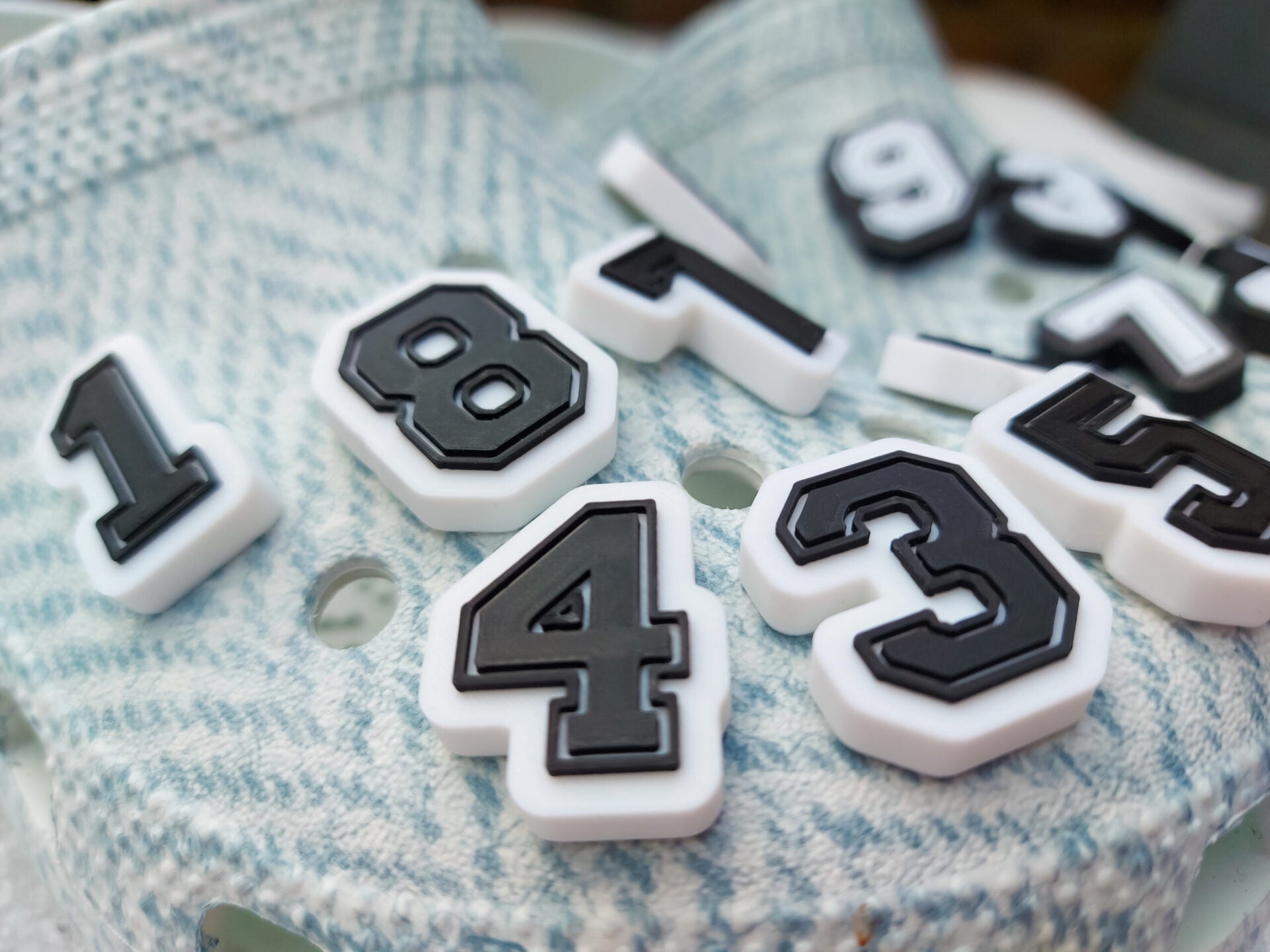 A pair of blue-patterned shoes featuring custom 3D-printed number charms, showcasing the digits 0 through 9 in sleek black and white colors. Each charm is securely clipped into the shoe holes, standing out with a bold varsity-inspired style.