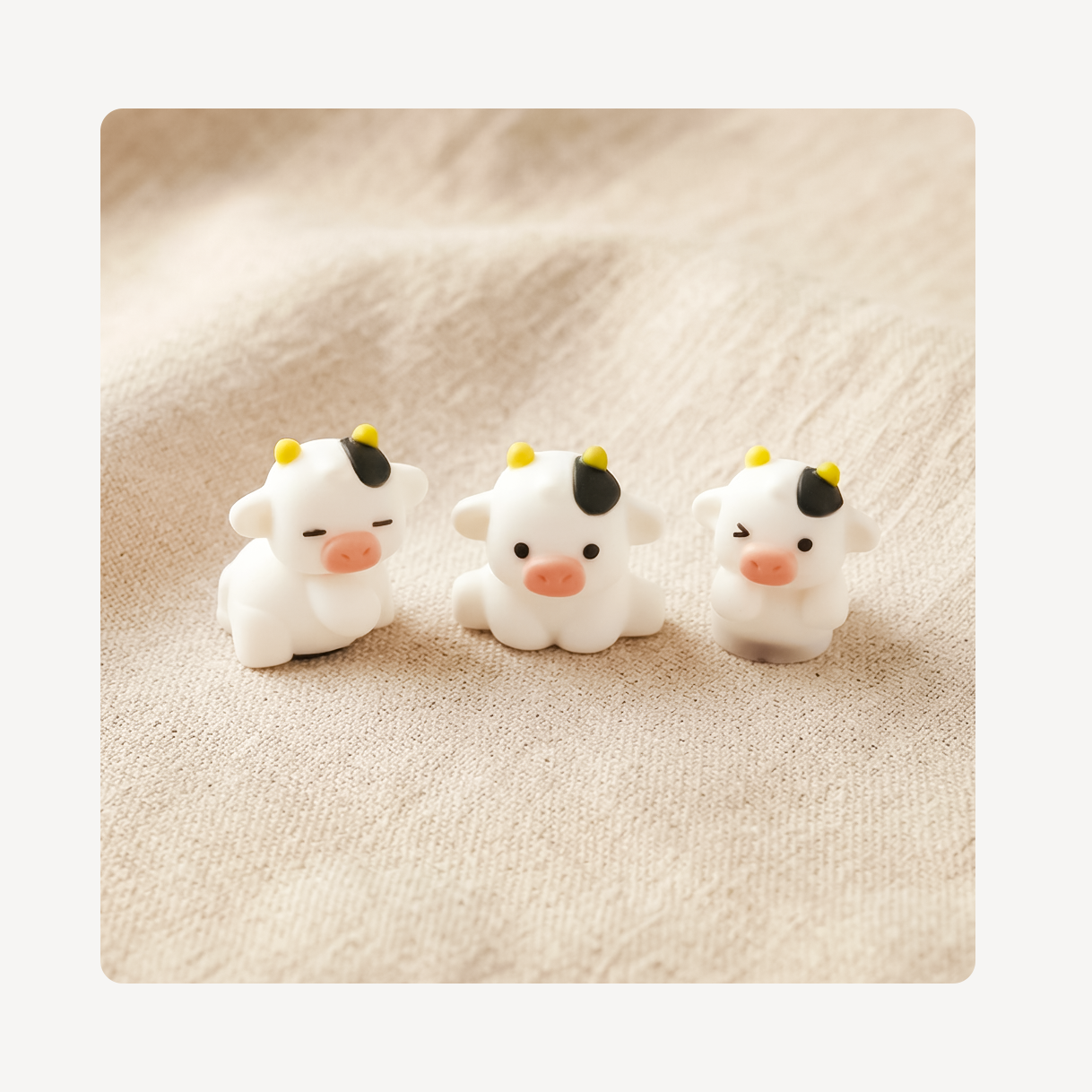 Three small white cow clog charms with black and yellow details, arranged side by side on a beige textured fabric background.