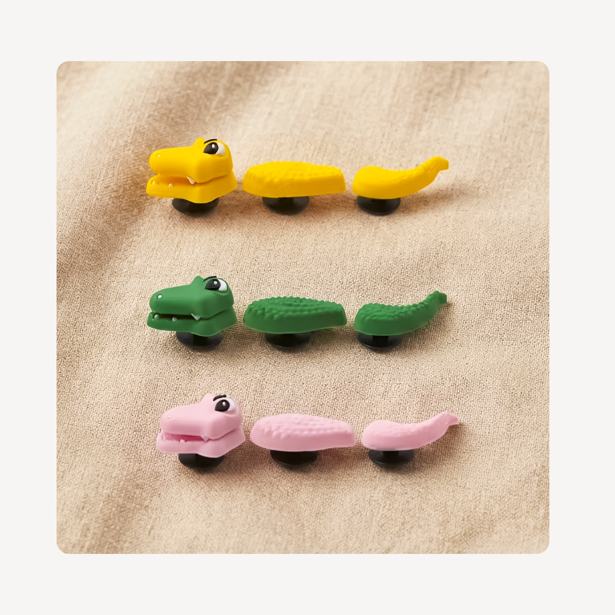 Three pairs of colorful alligator-shaped clog charms in yellow, green, and pink, arranged horizontally on a textured beige fabric background.