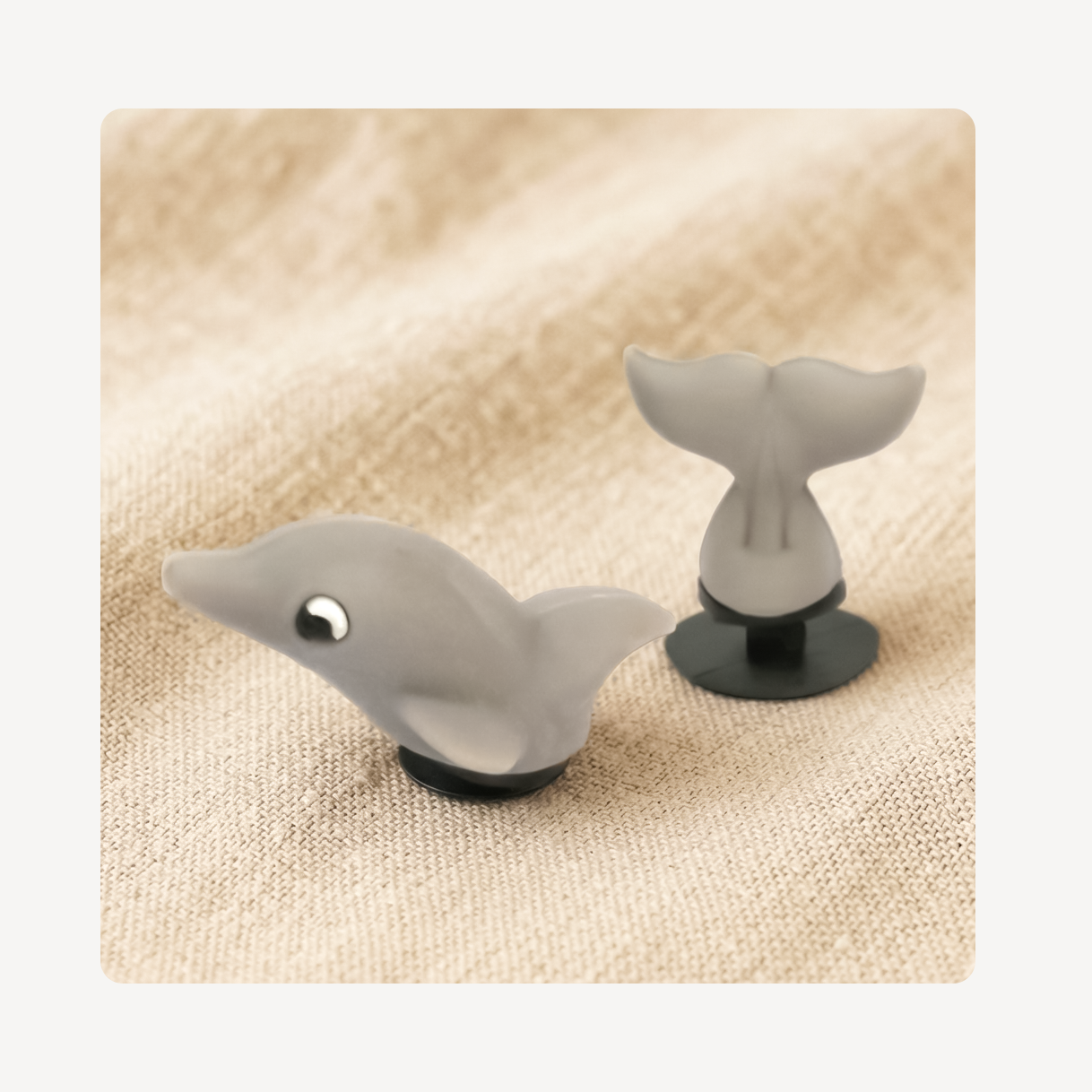 A pair of small gray clog charms shaped like a dolphin with a tail, displayed on a textured beige fabric background.
