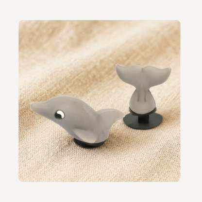 A pair of small gray clog charms shaped like a dolphin with a tail, displayed on a textured beige fabric background.