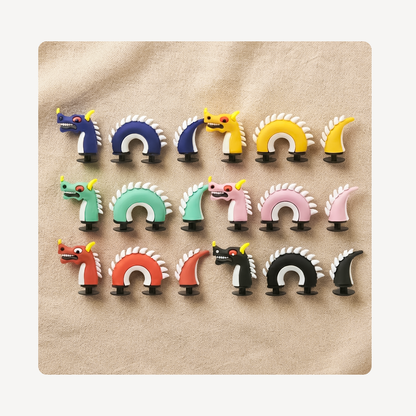 Six colorful dragon-shaped clog charms arranged in three rows on a beige textured background. The set includes blue, yellow, green, pink, red, black, and white versions, with some featuring small yellow horn accents.