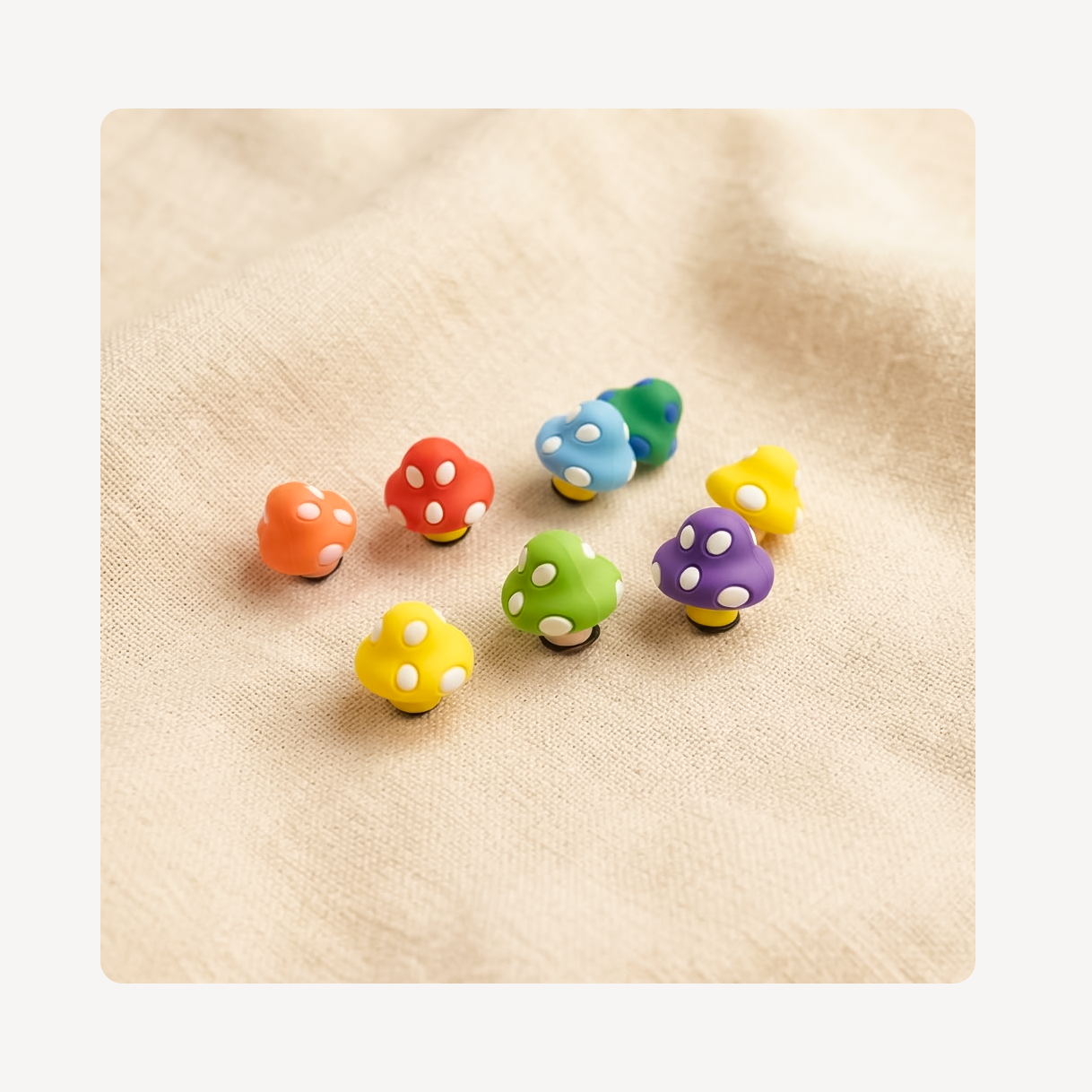 Seven small mushroom-shaped clog charms in assorted colors including orange, red, blue, green, yellow, purple, and teal, arranged on a beige textured fabric background.