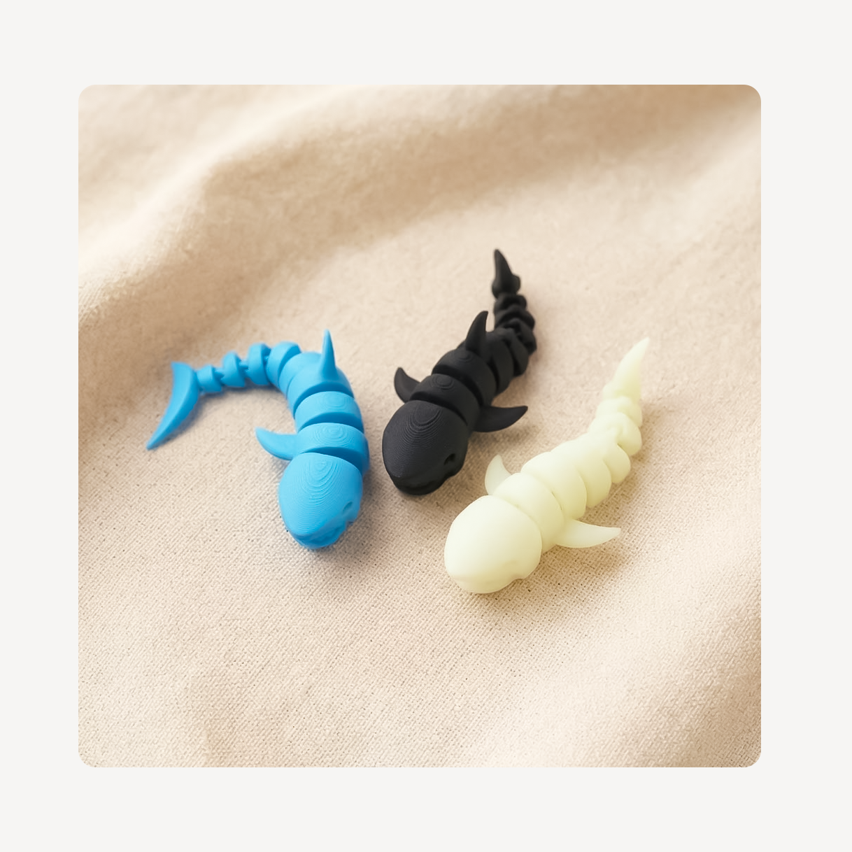 Three small shark-shaped clog charms in blue, black, and pale cream are arranged on a soft beige fabric background.