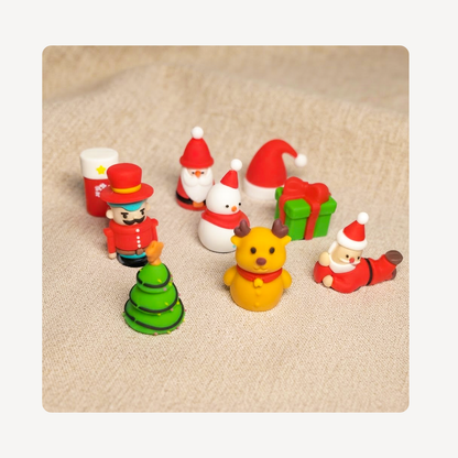 A set of small Christmas-themed clog charms arranged on a beige textured fabric background. The set includes Santa Claus figures in red and white, a yellow bear-like character, a green Christmas tree, a snowman, a nutcracker, a red hat, and a green gift box with red ribbon.