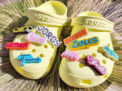 A pair of yellow shoes shoes featuring a variety of colorful, custom name charms. Each charm showcases different names in unique fonts. They allow you to personalize names or phrases with chosen colors and fonts, making each design unique.