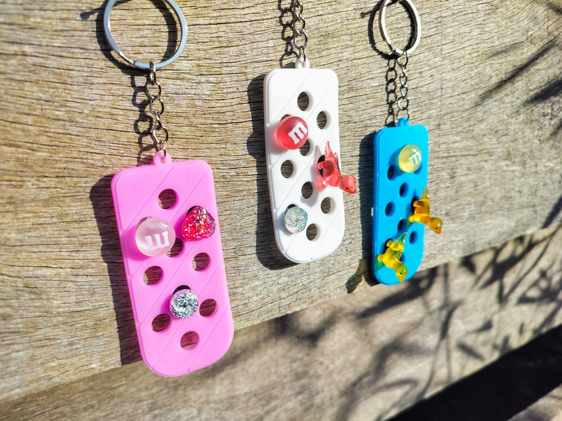 Three PVC keychains in pink, white, and blue with holes designed to hold shoe charms. Each keychain displays a unique set of charms, including candy-themed and colorful glitter charms, showcasing a playful and customizable accessory for charm lovers.