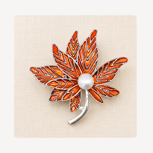 An autumn maple leaf clog charm featuring vibrant orange and red enamel, a white faux-pearl center, and a silver-tone metal outline and stem, displayed on a textured beige background.