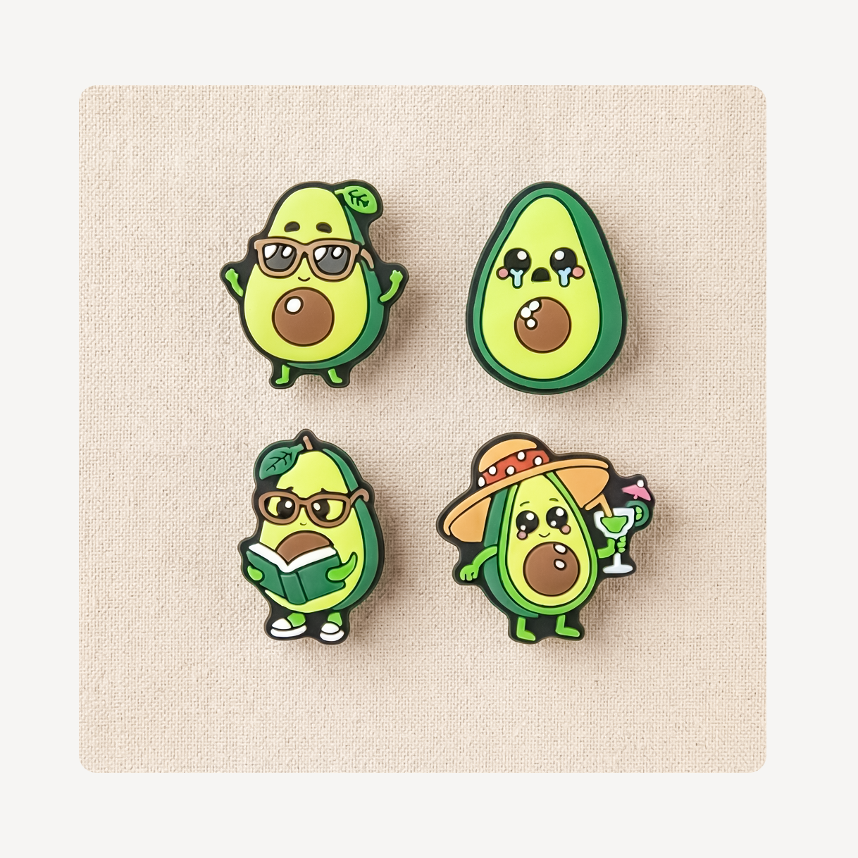 Four cute avocado character clog charms arranged in a 2x2 grid on a beige textured square background. The avocados have different expressions and accessories: one wears sunglasses and waves, one is crying, one reads a book, and one wears a straw hat and holds a cocktail glass.