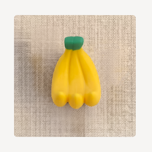 A yellow banana-shaped clog charm with a green top, centered on a textured beige fabric background.