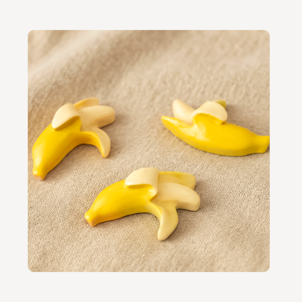 Three small banana-shaped clog charms in yellow and cream are arranged on a beige textured fabric background.