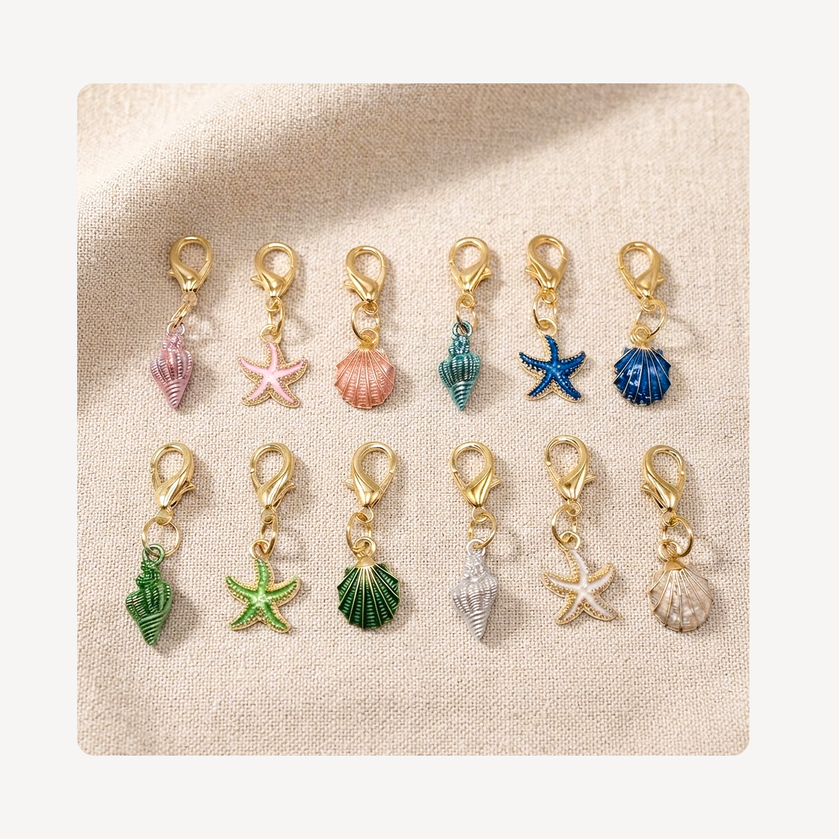 Twelve small gold-tone lobster clasp shoe charms arranged in two rows on a beige textured fabric background. The charms feature colorful seashell and starfish designs in pink, peach, teal, blue, green, white, and beige.