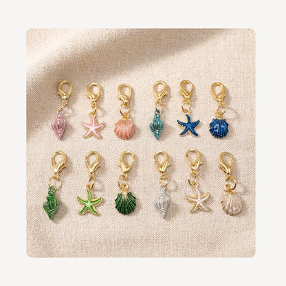 Twelve small gold-tone lobster clasp shoe charms arranged in two rows on a beige textured fabric background. The charms feature colorful seashell and starfish designs in pink, peach, teal, blue, green, white, and beige.