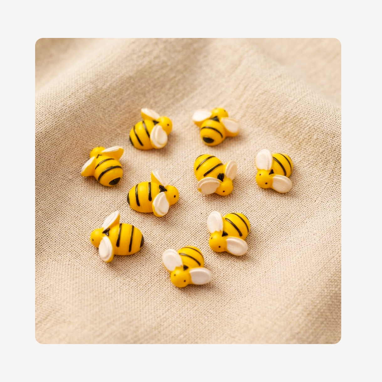 Nine small yellow and black striped bee-shaped clog charms with white wings are arranged on a beige textured fabric background.