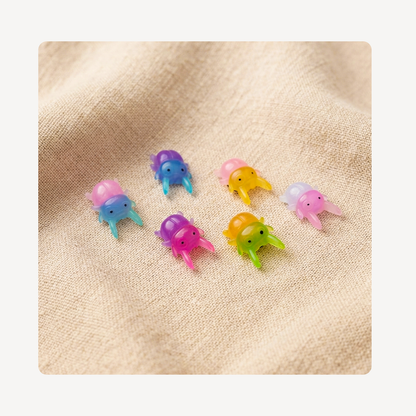 Six small beetle clog charms in assorted pastel and bright colors, arranged on a textured beige fabric background.