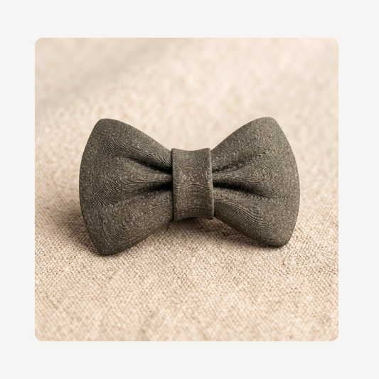 A small bow tie-shaped clog charm in a textured black plastic material, centered on a beige woven surface.