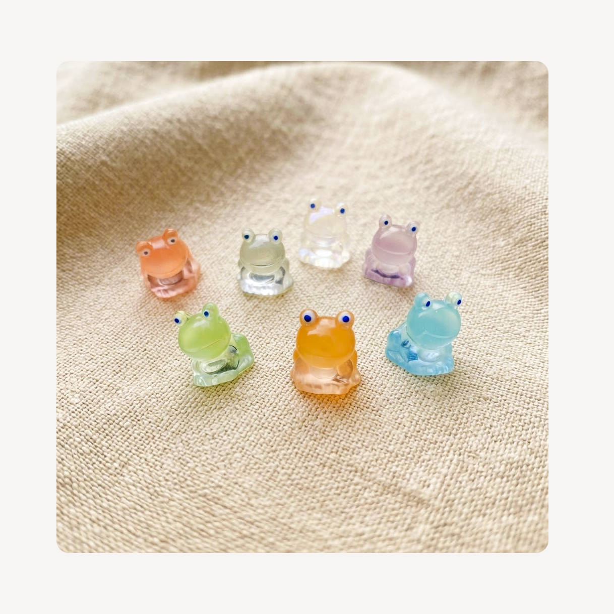 Seven small translucent frog-shaped clog charms in pastel colors, including peach, pale green, clear, lavender, mint green, and light blue, arranged on a beige textured fabric background.