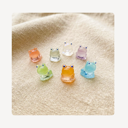 Seven small translucent frog-shaped clog charms in pastel colors, including peach, pale green, clear, lavender, mint green, and light blue, arranged on a beige textured fabric background.