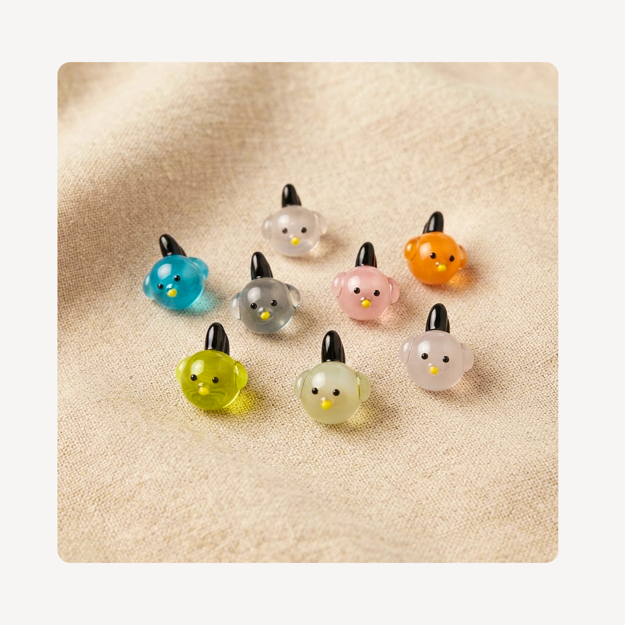 Eight small translucent bird-shaped clog charms in assorted colors, arranged on a soft beige fabric background.