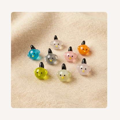 Eight small translucent bird-shaped clog charms in assorted colors, arranged on a soft beige fabric background.