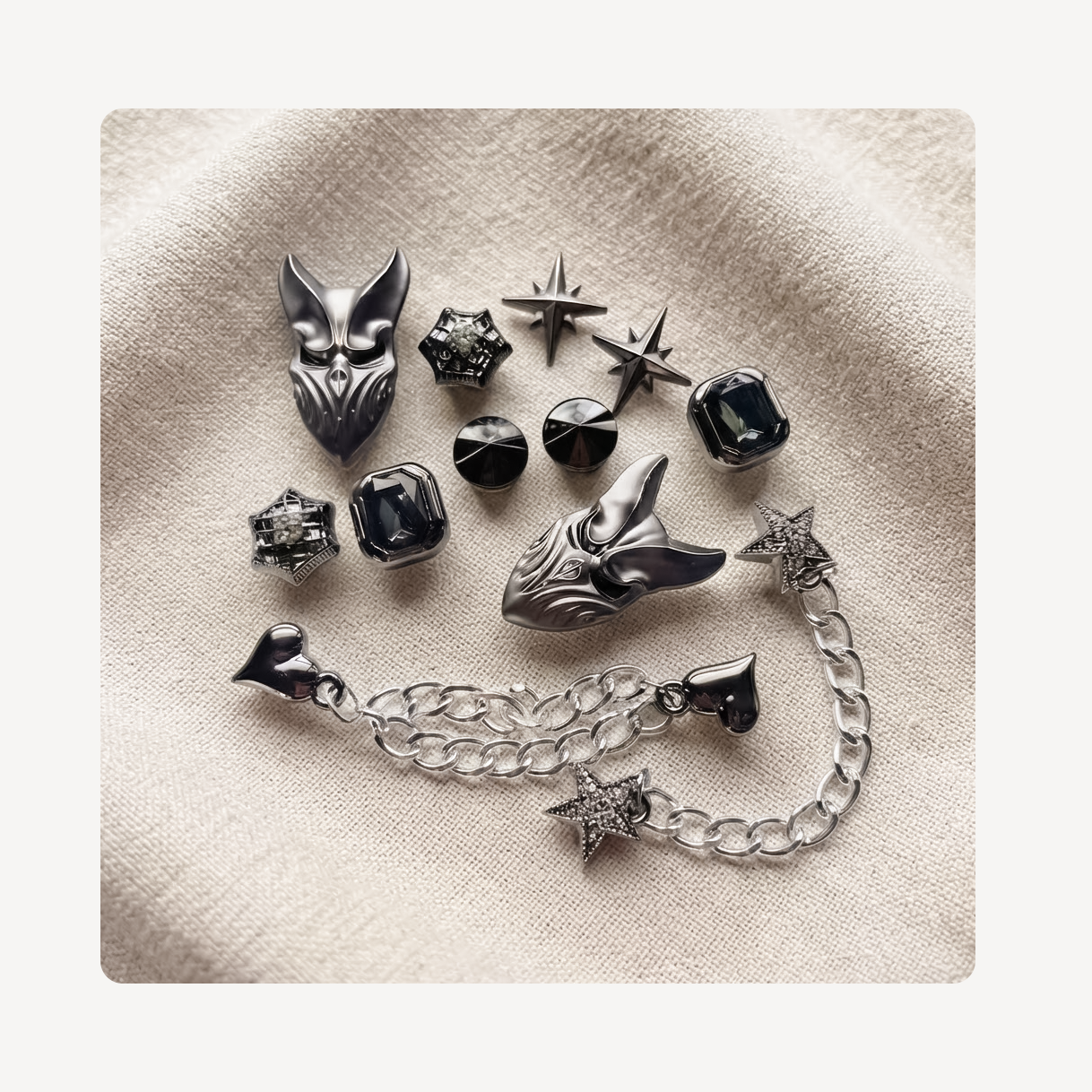 A flat-lay arrangement of gothic clog charms on a textured beige fabric background, featuring silver and black fox head charms, star-shaped earrings, faceted black gemstones, and a silver chain with heart and star charms.