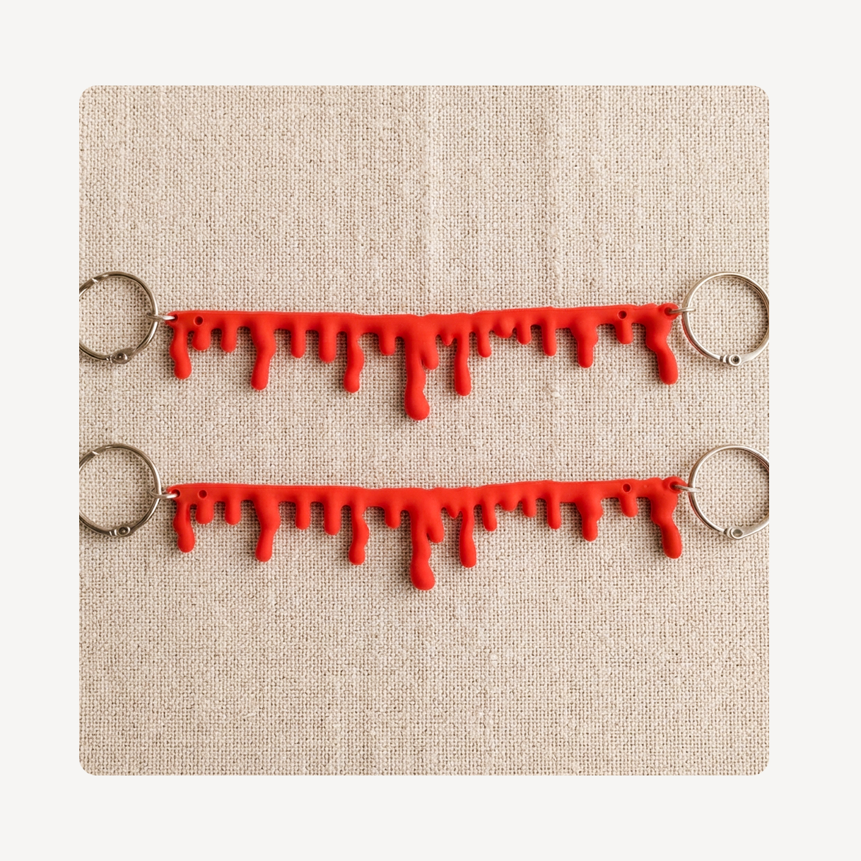 Two red dripping blood or paint clog chains with silver metal key rings, displayed on a textured beige linen background.