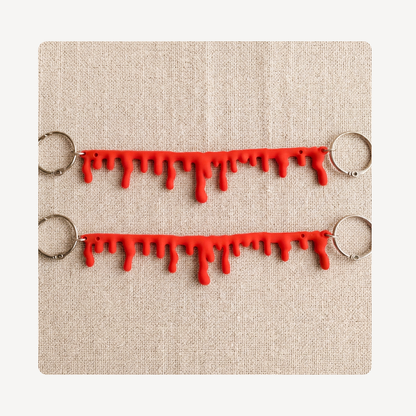 Two red dripping blood or paint clog chains with silver metal key rings, displayed on a textured beige linen background.