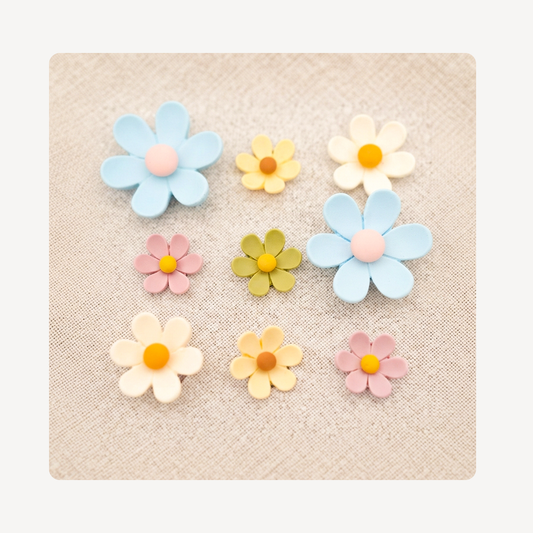 Nine pastel flower clog charms with yellow centers and pink, blue, and green petals, arranged on a textured beige fabric background.