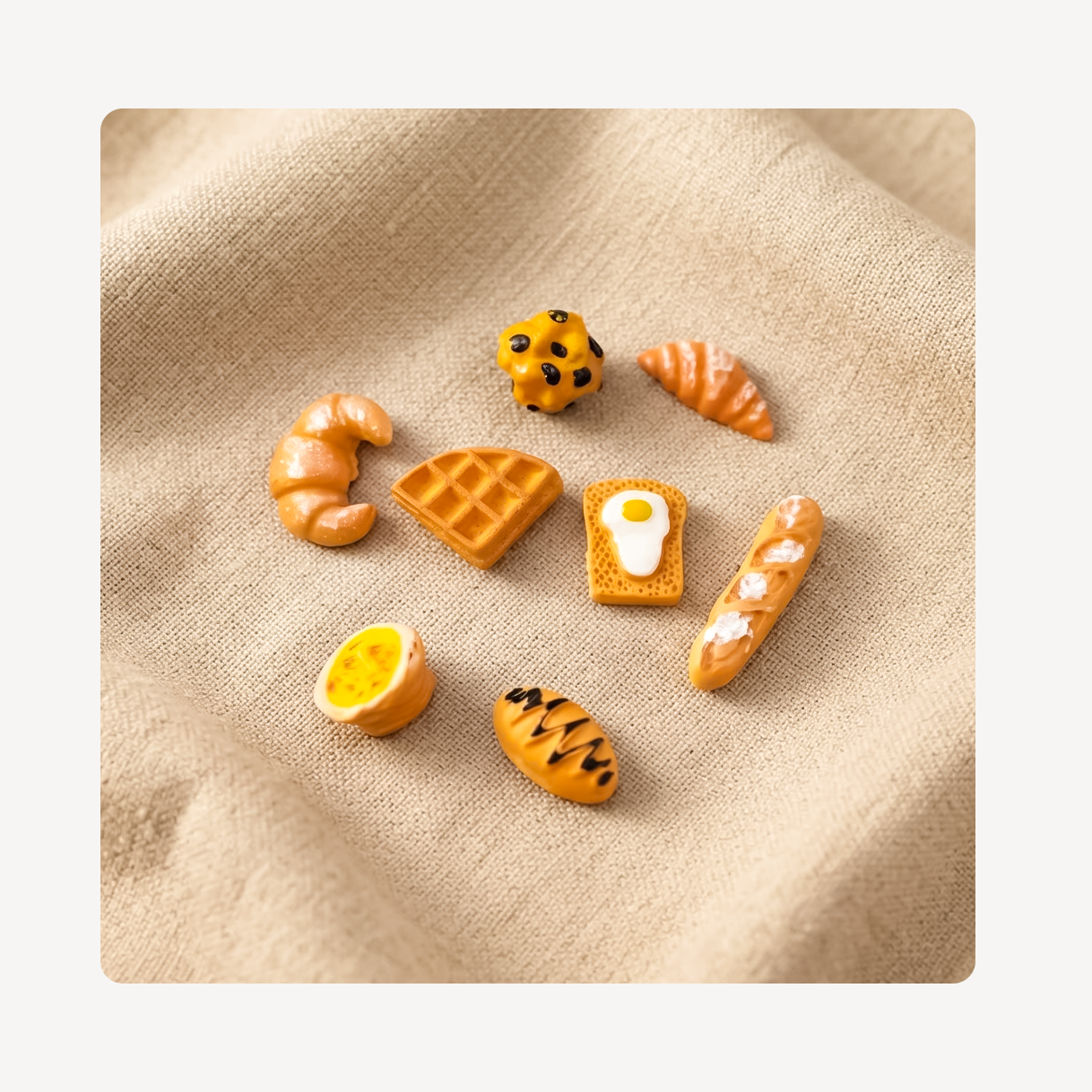 A set of miniature food-themed clog charms shaped like croissants, waffles, toast with an egg, a baguette, an orange slice, and a chocolate chip cookie, arranged on a beige textured fabric background.