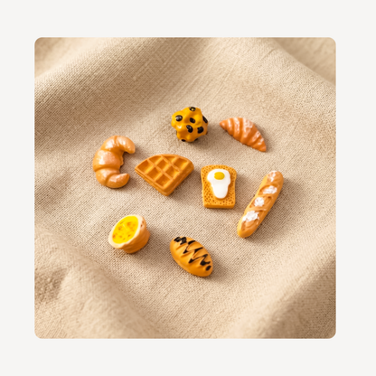 A set of miniature food-themed clog charms shaped like croissants, waffles, toast with an egg, a baguette, an orange slice, and a chocolate chip cookie, arranged on a beige textured fabric background.