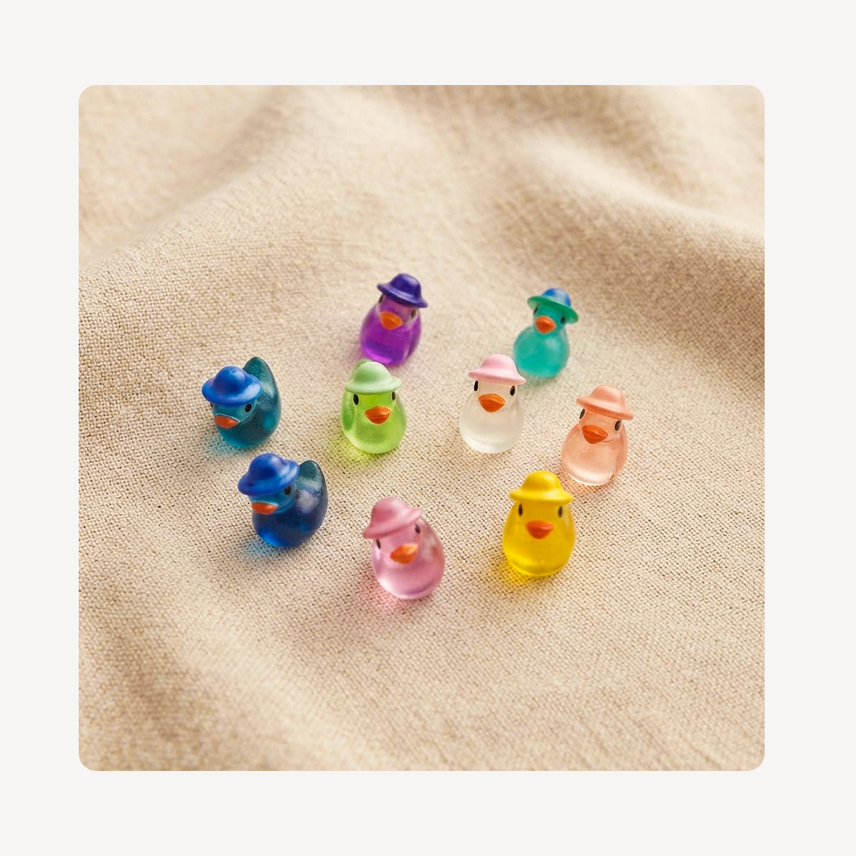 Nine small translucent duck clog charms in assorted colors, each wearing a different colored hat, arranged on a beige textured fabric background.