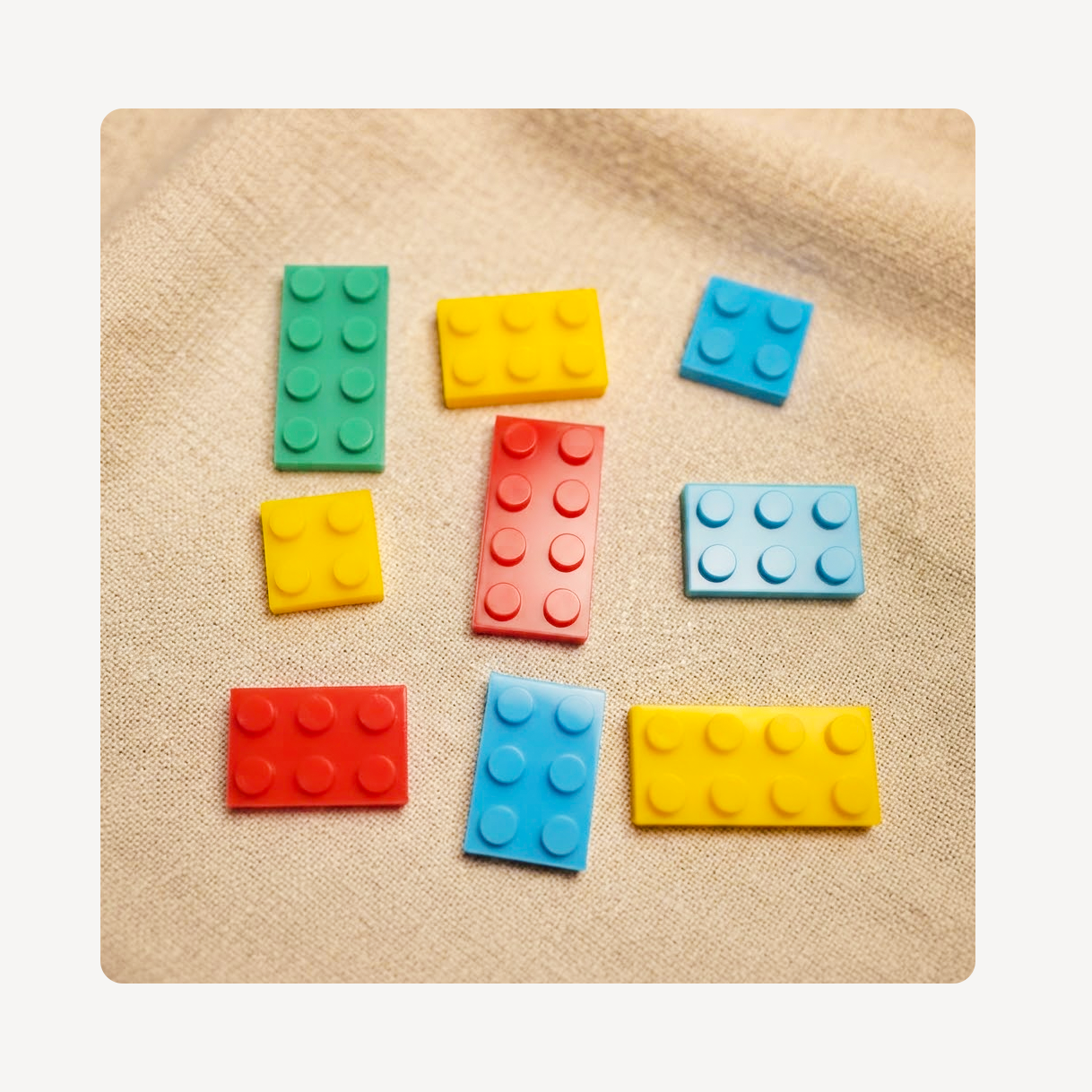 A set of colorful building block clog charms arranged on a beige textured fabric background. The charms come in green, yellow, blue, red, and light blue colors, with different sizes.