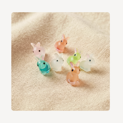 Seven small translucent bunny clog charms in pastel colors including pink, peach, teal, white, and light green, arranged on a textured beige fabric background.