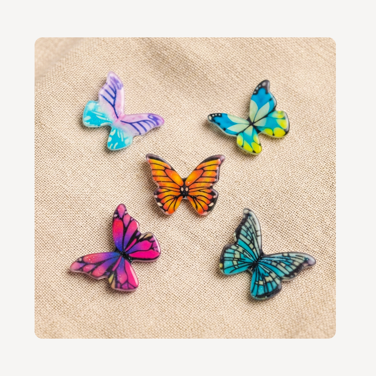 Five colorful butterfly-shaped clog charms arranged on a beige textured fabric background.