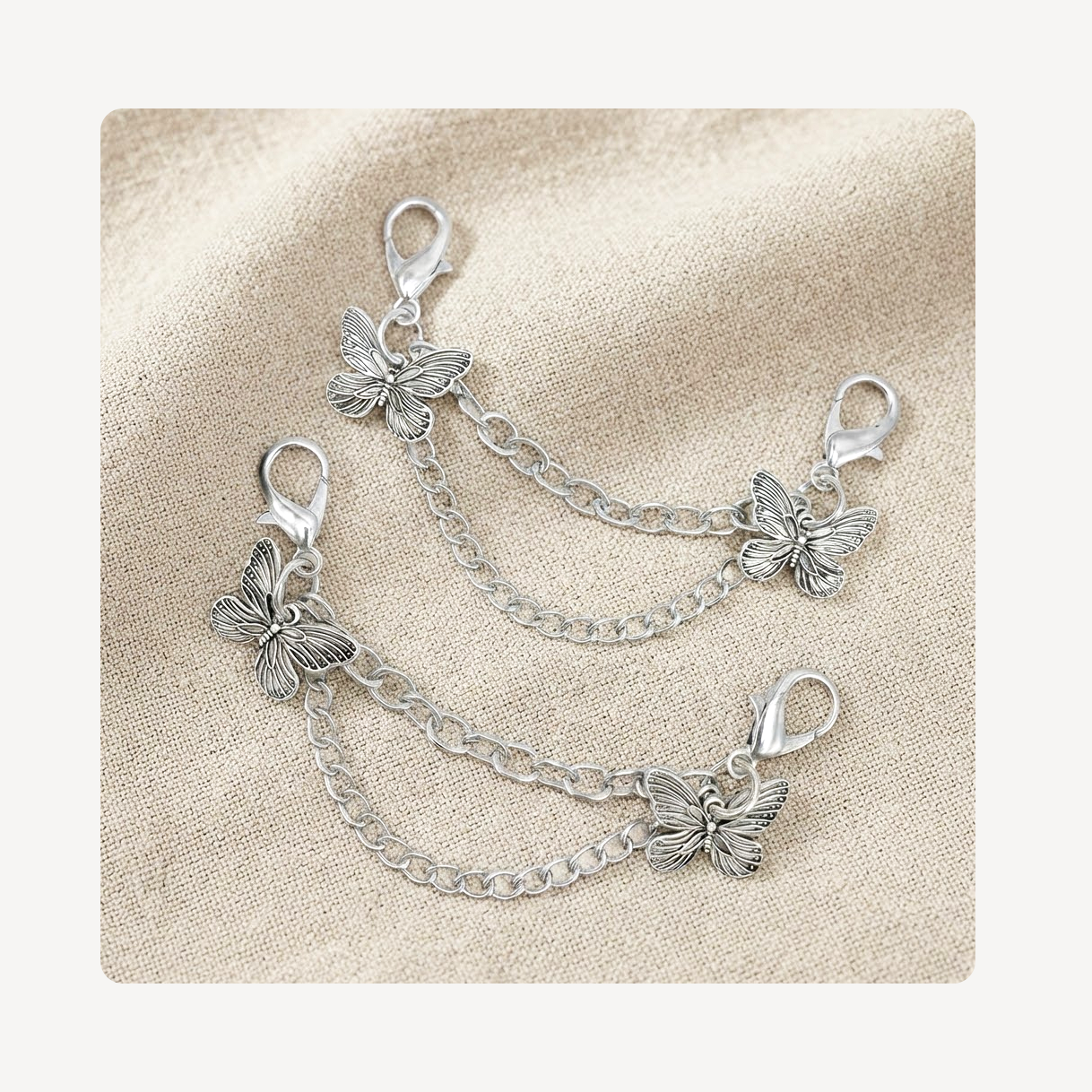 Two silver butterfly shoe chain link charms with lobster clasp ends, arranged on a textured beige fabric background.