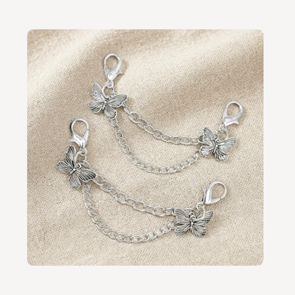 Two silver butterfly shoe chain link charms with lobster clasp ends, arranged on a textured beige fabric background.