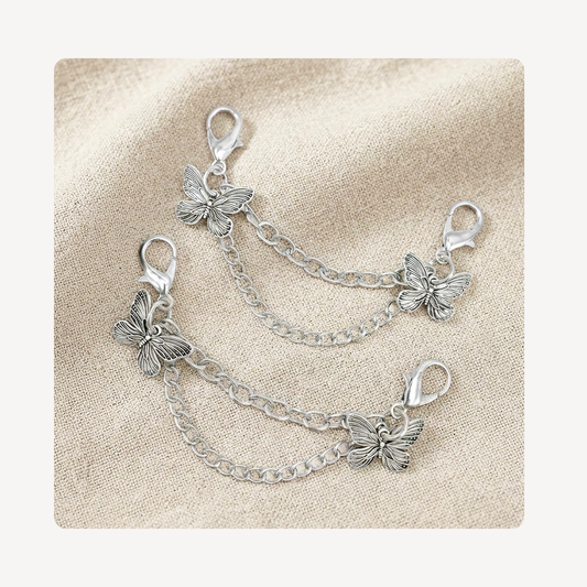 Two silver butterfly shoe chain link charms with lobster clasp ends, arranged on a textured beige fabric background.