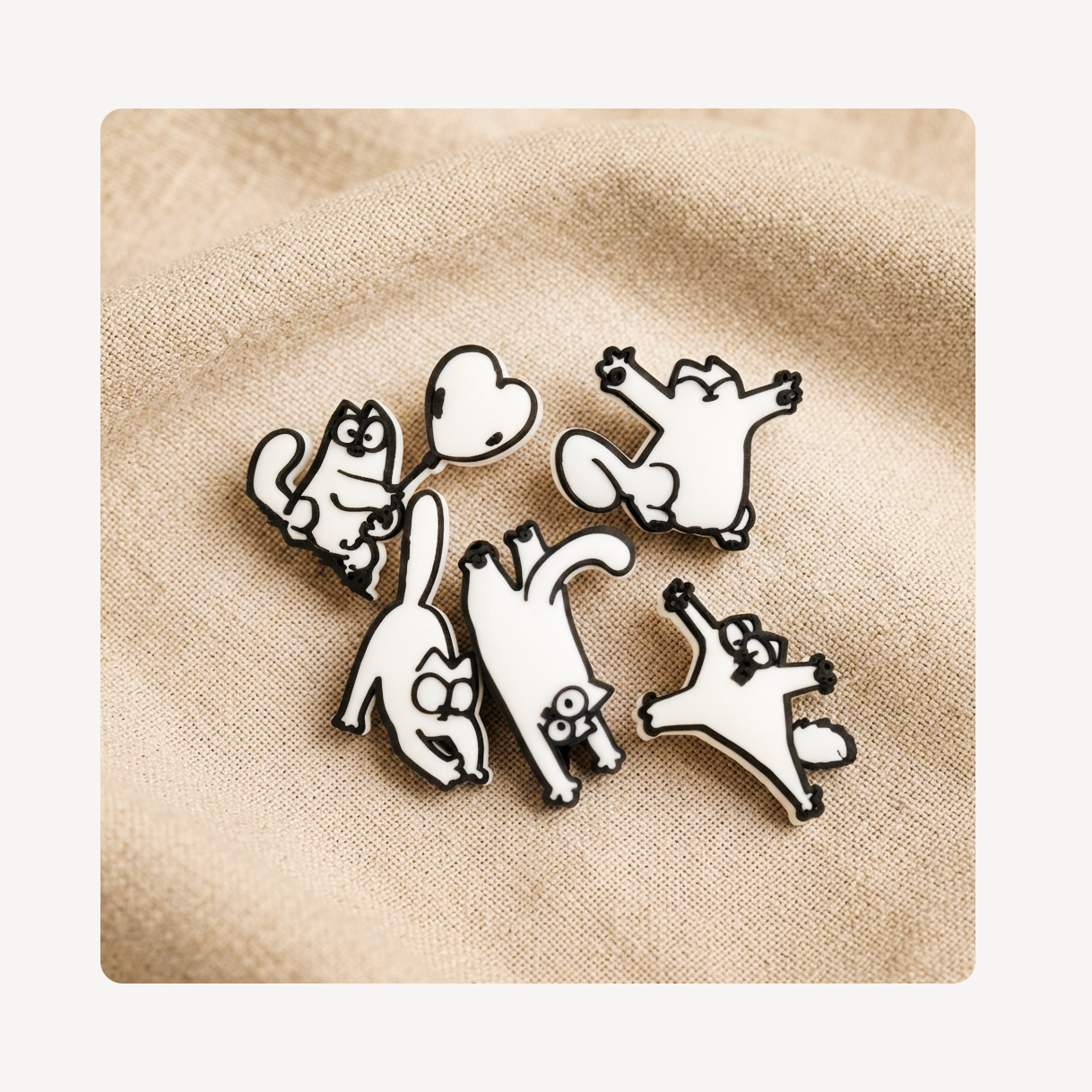 Five white cartoon cat clog charms with black outlines, arranged on a beige textured fabric background.