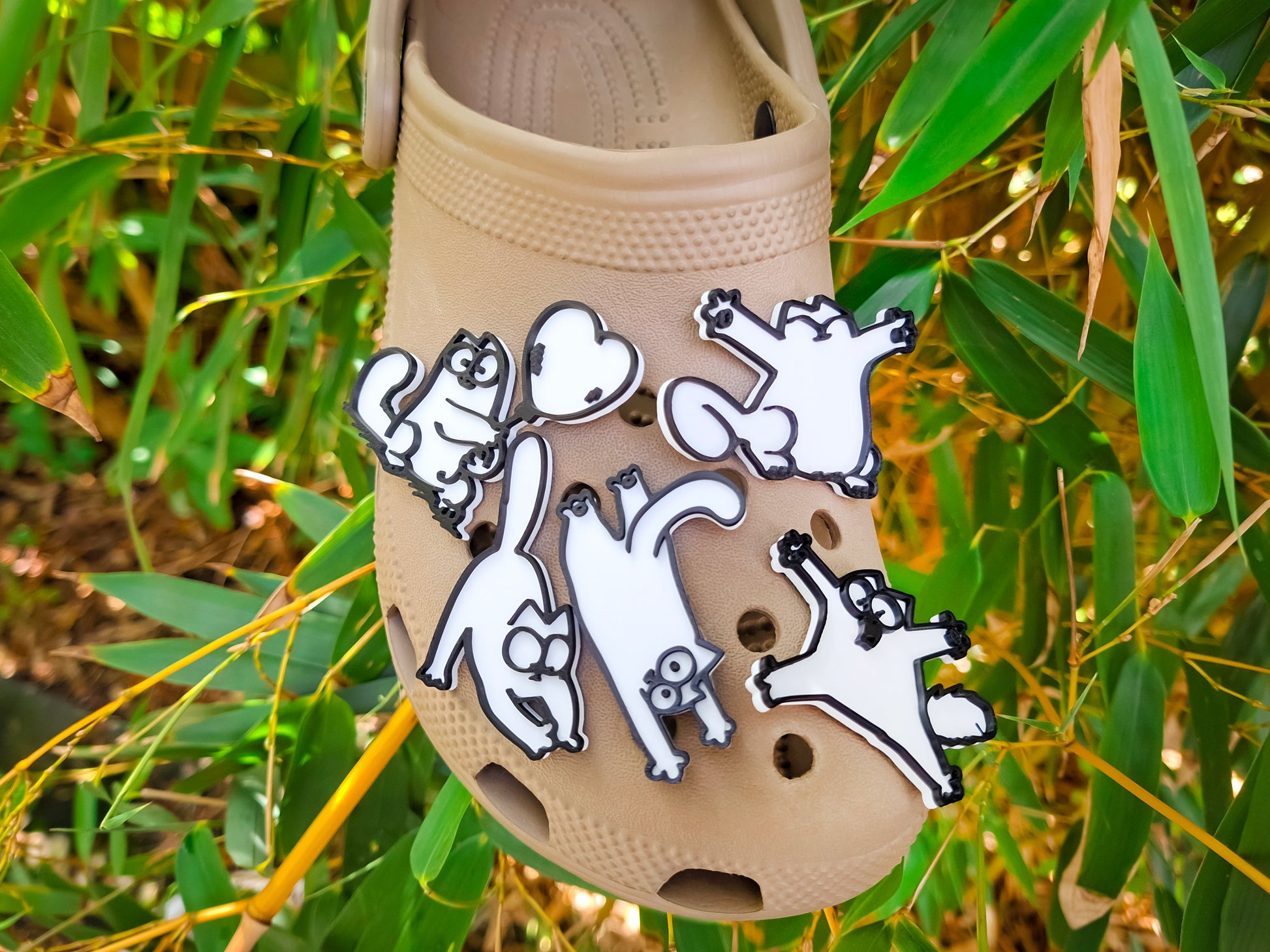 A beige Croc-style clog shoe decorated with five white cartoon cat clog charms outlined in black, photographed outdoors against a background of green bamboo leaves.